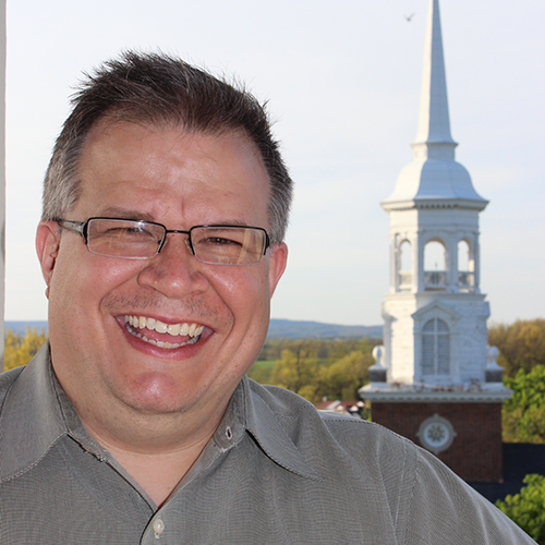 Shuan is wearing a gray shirt with a church steeple in the background